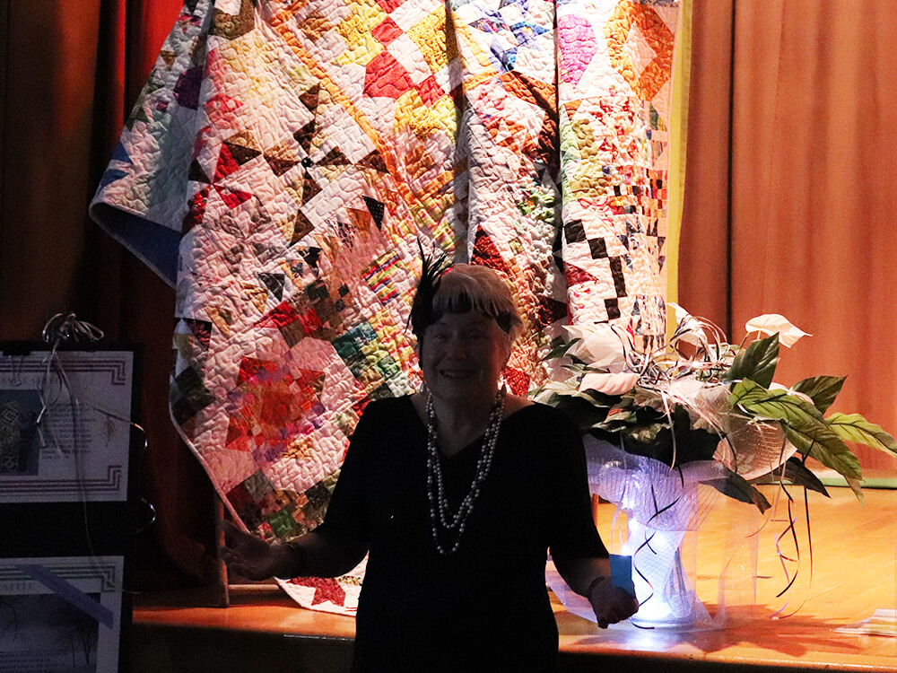 Picture of patron at the 20th-anniversary gala posing in front of a quilt that was raffled at the event